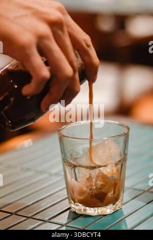Der Barista gießt frischen Espresso in ein Glas mit Eiswürfeln auf der Kaffeetheke und schließt die Zubereitung von Eiskaffee aus Stockfoto