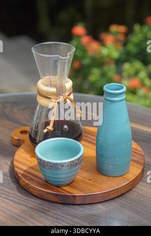 Manuelles Gießen über Kaffee gebrüht in Glaskaraffe mit Keramikbecher und -Flasche auf Holzbrett, natürlicher Hintergrund im Freien Stockfoto