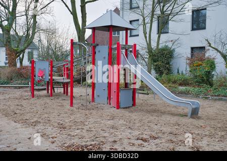 Verlassene graue und rote Spielplatzstruktur mit Rutschkletterwand und Dach in Sandy-Gegend inmitten von Bäumen und modernen Wohngebäuden im Vorort Sett Stockfoto