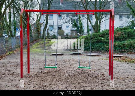Leere Red Frame Schaukeln mit grünen Sitzen auf Sandy Playground umgeben von Bäumen und Wohngebäuden in ruhiger Vorstadt Park Gegend Stockfoto