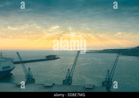Industriehafenkrane und ein großes Schiff stehen vor einem dramatischen Sonnenuntergang, bei dem Sonnenstrahlen durch die Wolken über dem Meer brechen Stockfoto