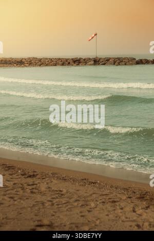 Eine ruhige Strandszene bei Sonnenuntergang mit sanften Wellen, die auf das Ufer Rollen, einem felsigen Steg in der Ferne und einer rot-weißen Flagge an einem Pol gegen ein W Stockfoto