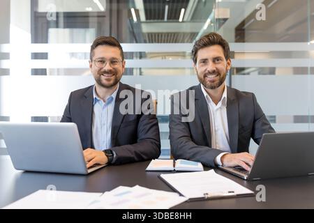 Zwei lächelnde Geschäftskollegen sitzen an einem Schreibtisch, blicken auf den Zuschauer und arbeiten während eines Firmenmeetings in einer hellen, modernen Büroumgebung an Laptops Stockfoto