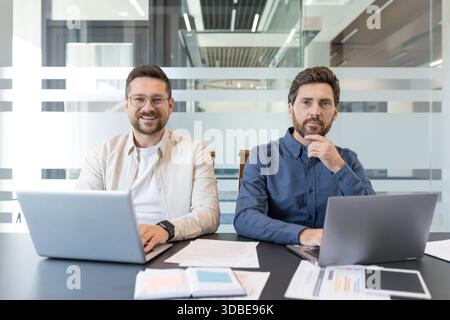 Zwei professionelle Geschäftsleute, die lächelnd an einem Konferenztisch mit Laptops sitzen und produktiv in einer modernen Büroumgebung zusammenarbeiten Stockfoto