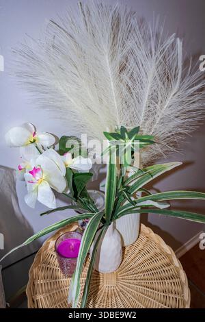 Stilvolle Innenausstattung mit einer weißen Vase mit künstlichen Orchideen, gestreiften Blättern und Federfedern, gepaart mit einer lila Kerze. Stockfoto