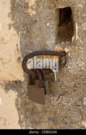 Detail der abgelösten Wand eines Gebäudes mit einem Loch in der Wand. Eine rostige Schlinge und drei alte Vorhängeschlösser. Stockfoto