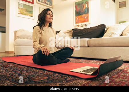 Frau mittleren Alters übt Achtsamkeit und Entspannung zu Hause während einer Arbeitspause, sitzt auf einer Yogamatte mit einem Laptop, Konzept der Work Life Balance Stockfoto