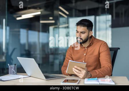 Fokussierter bärtiger junger Mitarbeiter am Schreibtisch, der während eines virtuellen Meetings in einem modernen Büro an einem Laptop arbeitet und Notizen schreibt, Aufgaben plant und organisiert Stockfoto