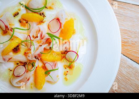Fisch-Ceviche liegt auf einer weißen Keramikplatte, garniert mit Orangensegmenten, Rettichen und grünen Zwiebeln. Ein glitzerndes Öl verstärkt die Farben dieses A Stockfoto