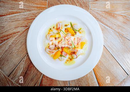 Auf einem runden Teller werden frische Fisch-Ceviche mit durchscheinenden Meeresfrüchten, Zitrussegmenten und dünn geschnittenen Radieschen sowie Microgreens und ein gl gezeigt Stockfoto