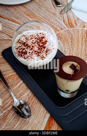 Zwei klare Glasbecher zeigen Tiramisu-Desserts, die mit dunkler Schokolade und Kakaopulver auf Schlagsahne bestäubt sind. Ein Löffel liegt neben den Desserts auf einem Stockfoto