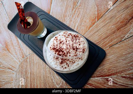 Zwei Tiramisu-Desserts, einer in einem kurzen Glas mit dunkelbrauner Flüssigkeit und der andere in einem mit Gewürzen bestäubten Glas, ruhen auf einem schwarzen Tablett auf einer Woo Stockfoto