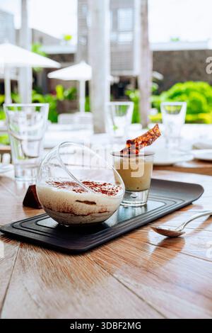 Eine klare Glasschale und eine kleine Tasse zeigen Tiramisu-Dessert mit Schokoladensplittern auf einem schwarzen Serviertablett auf einem Holztisch. Wassergläser Stockfoto