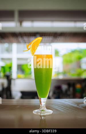 Ein lebendiges mehrschichtiges Getränk befindet sich in einem klaren Glas, das mit einer Orangenschale garniert ist, auf einer dunklen Holzplatte, eingerahmt von Laub. Stockfoto