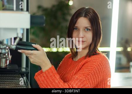 Rückansicht eines brünetten Barista in Strickmode, der Kaffee mit einem sanften Lächeln in gemütlicher Inneneinrichtung zubereitet. Stockfoto