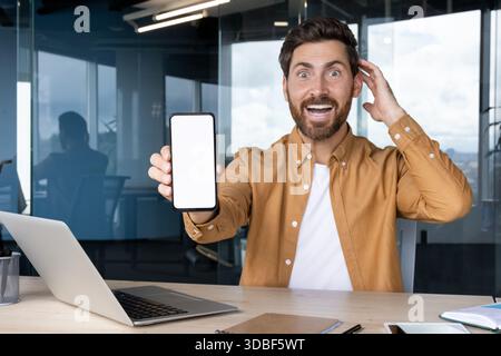 Ein aufgeregter Mann sitzt an einem Schreibtisch, hält ein Smartphone mit einem leeren weißen Bildschirm für Inhalte in der Hand, der Überraschung und Glück mit einem offenen Mund und einer Hand ausdrückt, die seinen Kopf berührt Stockfoto