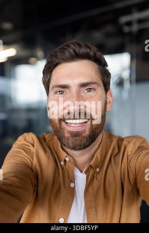 Bärtiger Mann lächelt und blickt in die Kamera, macht Videoanrufe oder macht ein Selfie in einer Büroumgebung, vermittelt Konzepte von Selbstvertrauen, Verbindung und Fernkommunikation Stockfoto