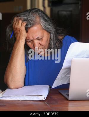 Eine ältere Latina sitzt zu Hause an einem Tisch, liest Dokumente, benutzt einen Laptop und schreibt Notizen mit einem fokussierten, seriösen Ausdruck in ein Notizbuch Stockfoto