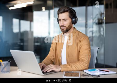 Konzentrierter Mann, der schnurlose Kopfhörer trägt, an einem Schreibtisch sitzt und auf einem Laptop schreibt, widmet sich seiner Arbeit in einer modernen Büroumgebung Stockfoto