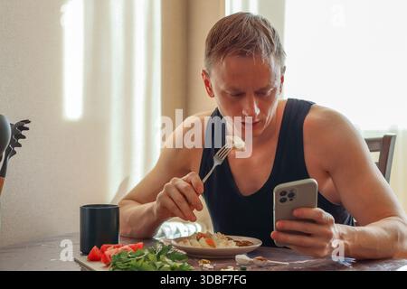 Sportlicher Mann im ärmellosen Top Check-Gerät, hebt Lebensmittel auf Gabel aus dem Gericht, zwischen Gemüse und Nahrungsergänzungsmitteln Stockfoto