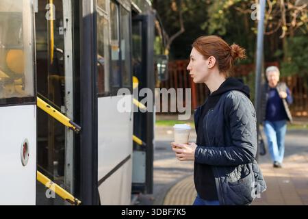 Zwanglos gekleidete Frau hält Kaffee, nähert sich der offenen Bustür mit verschwommenen Fußgängern in sonniger urbaner Umgebung. Stockfoto