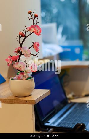 Künstliche rosafarbene Blüten in weißem Topf auf einer hölzernen Schreibtischecke in der Nähe des Laptopbildschirms Stockfoto