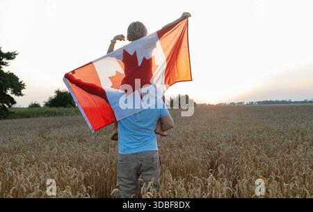 Große kanadische Flagge über dem Jungen, der auf den Schultern des Menschen sitzt und bei Sonnenuntergang in einem Weizenfeld steht. Happy Canada Day Stolz, Freiheit, Patriotismus. A j Stockfoto