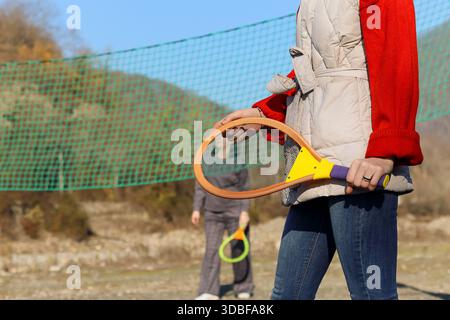 Fokussierte Aufnahme einer weiblichen Hand, die einen lebhaften Schläger im Vordergrund hält, mit verschwommenen Tennisspielern auf dem Platz in sonniger Umgebung. Stockfoto