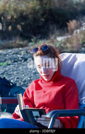 Legere Frau mit Pferdeschwanz und Sonnenbrille, in rotem Pullover, sitzt auf dem Stuhl und liest Buch in felsiger Natur nahe dem Wasser Stockfoto