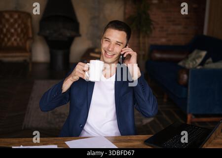 Selbstbewusster Mann, der auf dem Schreibtisch sitzt und sich von der Arbeit mit elektronischen Dokumenten auf dem Laptop absetzt, um einen Anruf entgegenzunehmen. Lächelnder junger Freelancer, der Kaffee aus der Tasse trinkt, während er am Telefon auf der Arbeit telefoniert || Model veröffentlicht Stockfoto