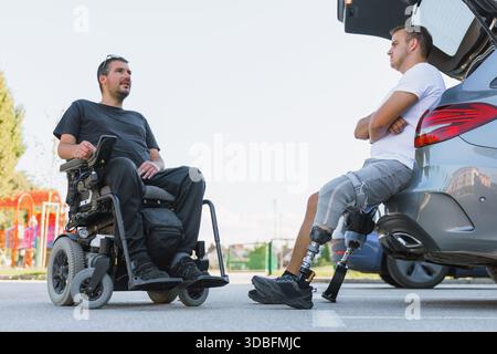 Zwei behinderte Freunde unterhalten sich in der Nähe eines Autos auf einem Parkplatz, einer auf einem Rollstuhl und einer mit einer Beinprothese. Stockfoto