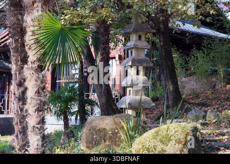 Zen-Garten mit hoher Steinlaterne neben dem Pavillon mit geschwungenem Dach, üppigen Palmen, hohem Bambus, moosigen großen Felsen und Herbstlaub unter sanftem Licht. Stockfoto