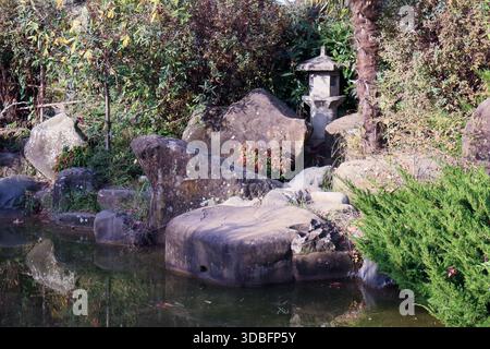 Traditionelle Steinlaterne in der Nähe eines reflektierenden Teichs mit moosbedeckten Felsen, leuchtenden roten Blumen, Palmen und dichten grünen Sträuchern in einer ruhigen Atmosphäre Stockfoto