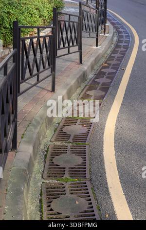 Konzentrieren Sie sich auf verwitterte Metallabflüsse, die in Betonkränze neben der gewundenen Straße eingebettet sind, mit schwarzem Eisenzaun und grünen Sträuchern darüber. Stockfoto