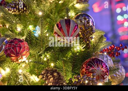 Nahaufnahme eines geschmückten Weihnachtsbaums mit leuchtenden Lichtern, roten und goldenen Ornamenten, Tannenzapfen und festlichen Feiertagsdetails vor den Gebäuden. Stockfoto