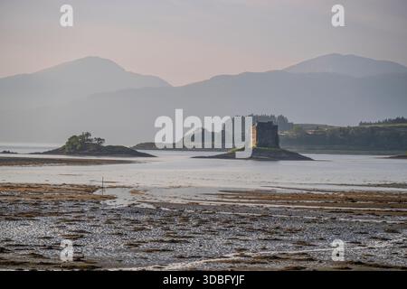 Eine ruhige Küstenszene mit einer einsamen Steinburg auf einer kleinen Insel, mit nebligen Bergen in der Ferne, ruhigem Wasser und einem reflektierenden Ufer Stockfoto