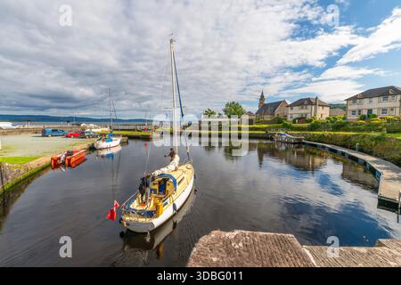 Ein kleines Segelboot ruht in einer von Steinen gesäumten Kanalschleuse am Ardrishaig Basin am Crinan Canal mit Booten, geparkten Autos und einer bezaubernden Skyline des Dorfes Stockfoto