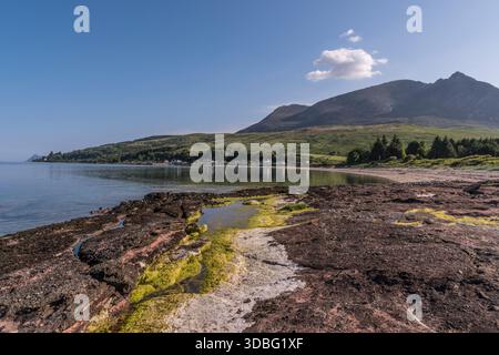 Ein ruhiges Küstenpanorama mit einer moosigen, felsigen Küste, ruhigem Wasser und sanften grünen Hügeln, die zu zerklüfteten Bergen unter einem hellblauen Himmel führen. Stockfoto