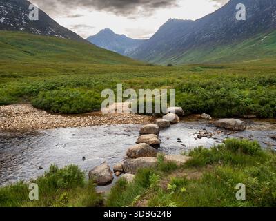 Eine ruhige schottische Landschaft mit einem flachen Fluss mit Trittsteinüberquerung, üppigen grünen Ufern und rauer Bergkulisse, die Ruhe vermittelt, o Stockfoto