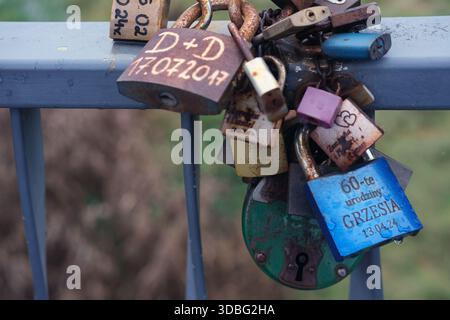 Nahaufnahme von farbenfrohen Vorhängeschlössern, die an einem horizontalen Metallgeländer befestigt sind, mit rostigen Ketten, sichtbaren Namen und Daten, natürlichem Tageslicht. Stockfoto