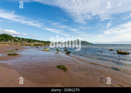 Eine ruhige Küstenszene in der Whiting Bay mit Sandstrand, moosigen Felsen, sanften Wellen und fernen grünen Hügeln unter einem hellblauen Himmel, perfekt für Stockfoto