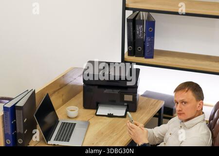 Geschäftsmann sitzt auf einem Stuhl am Schreibtisch und schaut sich in einem organisierten Bürobereich ein Smartphone mit Drucker, Laptop und Kaffeetasse an. Stockfoto