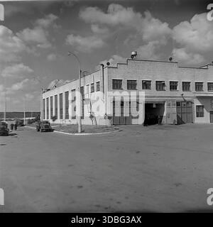 Schwarzweiß-Archivfoto eines sowjetischen Motordepots (autobaza) oder einer industriellen Garage in der Region Donezk, Ukraine, Donbass, 1960er Jahre Das weiße Backsteingebäude verfügt über große Reparaturbuchten und ein Metallsternsymbol auf dem Dach. Im Vordergrund steht eine alte GAZ-21 Volga Limousine. Lkw fahren im Hintergrund unter einem bewölkten Sommerhimmel entlang der Straße. Ein Blick auf die industrielle Infrastruktur und die Transportlogistik in der friedlichen Vorkriegszeit. Stockfoto