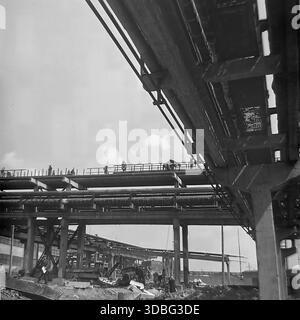 Schwarzweiß-Archivfoto, das eine riesige Industriebau im Donbass, Ukraine, UdSSR, 1960er Jahre zeigt Ein Niedrigwinkelfoto zeigt ein komplexes Netz von obenliegenden Rohrleitungen und Betonböcken, die sich darüber erheben. Silhouetten von Arbeitern sind sichtbar, die auf den hohen Gantry gegen den Himmel gehen. Bauschutt und eine Vintage-Lkw-Kabine liegen unten auf dem Boden. Ein starkes Bild der sowjetischen Schwerindustrie und Infrastrukturentwicklung. Stockfoto