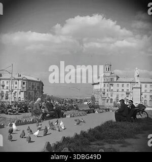 Schwarzweiß-Archivfoto, das eine friedliche Szene auf dem Hauptplatz von Druschkiwka, Region Donezk, Ukraine, Donbass, 1960er-1970er-Jahre zeigt Rentner und Kinder sitzen auf Bänken, die Tauben füttern. Das architektonische Ensemble umfasst ein markantes Wohngebäude mit einem Turm (belvedere) und einer entfernten Leninstatue. Ein nostalgischer Blick auf die generationenübergreifende Freizeit und die ruhige Atmosphäre eines sowjetischen Stadtplatzes lange vor dem Krieg. Stockfoto