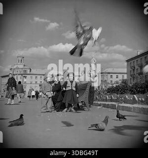 Schwarzweiß-Archivfoto, das eine friedliche Szene auf dem Hauptplatz von Druschkiwka, Region Donezk, Ukraine, Donbass, 1960er-1970er-Jahre zeigt Rentner und Kinder sitzen auf Bänken, die Tauben füttern. Das architektonische Ensemble umfasst ein markantes Wohngebäude mit einem Turm (belvedere) und einer entfernten Leninstatue. Ein nostalgischer Blick auf die generationenübergreifende Freizeit und die ruhige Atmosphäre eines sowjetischen Stadtplatzes lange vor dem Krieg. Stockfoto