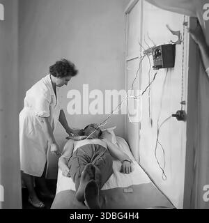 Schwarzweiß-Archivfoto, das eine Physiotherapie-Sitzung (Elektrotherapie) in einem sowjetischen Krankenhaus oder Sanatorium in der Donezk Region, Ukraine, Donbass, 1960er-1970er Jahre aufnimmt Eine Krankenschwester betreut einen männlichen Patienten, der auf einem Sofa liegt und über Kabel mit einem an der Wand montierten medizinischen Gerät verbunden ist. Das Verfahren umfasst wahrscheinlich Elektrophorese oder Galvanisierung. Ein ehrlicher Blick auf Gesundheitspraktiken und Rehabilitation in der UdSSR. Stockfoto