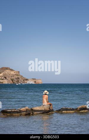 Eine Frau mit Sonnenhut sitzt auf Felsen am Meer in Elounda, Kreta, Griechenland, und blickt auf Spingalonga über dem ruhigen blauen Wasser. Stockfoto