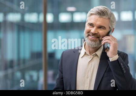 Reifer Geschäftsmann, der über ein Mobiltelefon kommuniziert, selbstbewusst lächelt und sich in einem professionellen Gespräch in einem Firmenbüro befindet und für Erfolg und Technologie steht Stockfoto