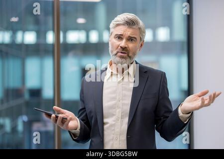 Reifer Geschäftsmann, der in einem modernen Büro steht, mit den Schultern zuckt und beide Hände offen hält, ein Mobiltelefon hält, Verwirrung, Unsicherheit oder Unwissenheit ausdrückt Stockfoto